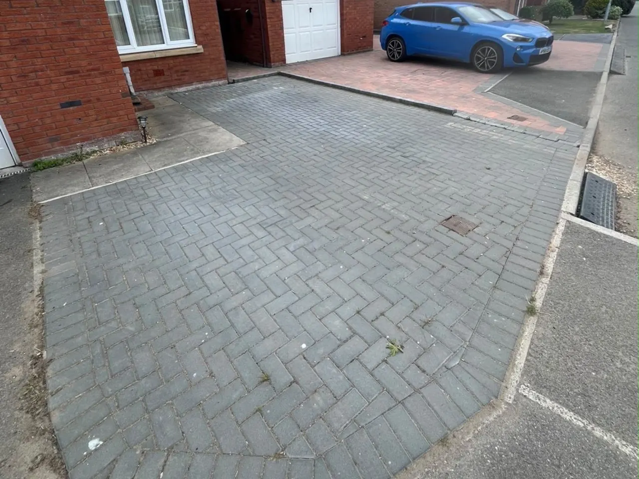 A block paving driveway before cleaning, the blocks dulled to a flat grey with weeds coming through the joints.